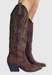 Bottes en daim marron jusqu'au genou avec des pointes effilées, ornées de détails métalliques en forme de flammes et d'un talon cubique texturé. Tirant décoratif sur la tige.