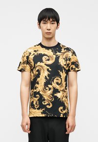Νέος άνδρας που φοράει μαύρο κοντομάνικο πουκάμισο με περίτεχνα χρυσά στροβιλιζόμενα μπαρόκ μοτίβα, στέκεται μπροστά σε απλό λευκό φόντο.