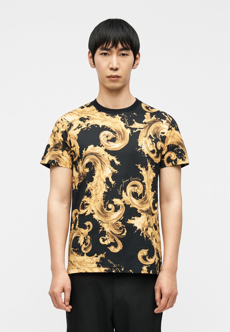 Νέος άνδρας που φοράει μαύρο κοντομάνικο πουκάμισο με περίτεχνα χρυσά στροβιλιζ όμενα μπαρόκ μοτίβα, στέκεται μπροστά σε απλό λευκό φόντο.