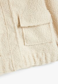 Cremefarvet, struktureret fleece-stof med en blød, fluffy finish. Har to påsyede lommer og lodrette syninger.