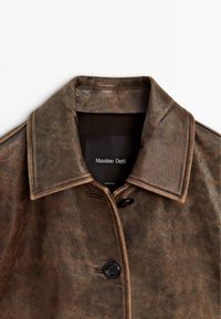 Giacca in pelle marrone con colletto strutturato, chiusura frontale a bottoni e texture liscia. L'etichetta riporta "Massimo Dutti". Finitura invecchiata visibile.