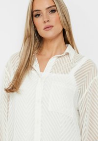 Leichte, durchsichtige weiße Bluse mit Chevron-Muster, Knopfleiste vorne und einer einzigen Brusttasche, versehen mit einem Kragen und langen Ärmeln.