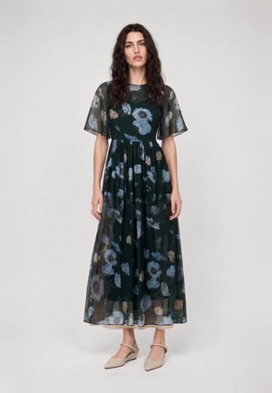 Frau trägt ein dunkles, durchsichtiges, geblümtes Maxikleid mit kurzen Ärmeln und beigen spitzen Schuhen und steht vor einem schlichten Hintergrund.