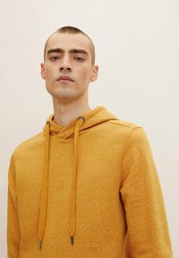 Sudadera amarilla mostaza hecha de tejido texturizado, con capucha ajustable, puños acanalados y un patrón sutil en toda su superficie.