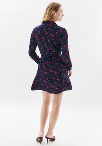 Kobieta w granatowej sukience w czerwony kwiatowy wzór, z przewiązanym paskiem w talii, długimi rękawami i brązowych szpilkach, zwrócona plecami na białym tle.