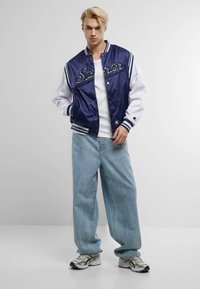 Navy and white varsity jacket with satin finish, featuring a "Starter" logo, striped ribbed cuffs and collar, paired with light blue baggy jeans and sneakers.