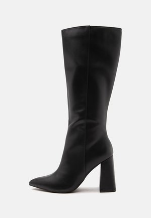 Botte haute en cuir noir avec un bout pointu et un talon haut épais sur un fond clair uni.