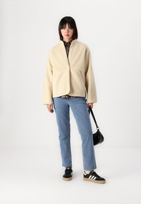 Beige fleecejakke med en avslappet passform, frontknapper og to lommer, kombinert med blå jeans og svarte joggesko med hvite striper.