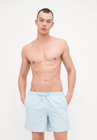 Lys blå badeshorts laget av hurtigtørkende stoff, med elastisk midjebånd og snøring samt sidelommer. Minimal merking synlig.