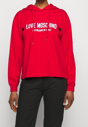 Personne portant un sweat à capuche rouge Love Moschino avec un texte blanc et des motifs de cœur noirs, associé à un pantalon noir.