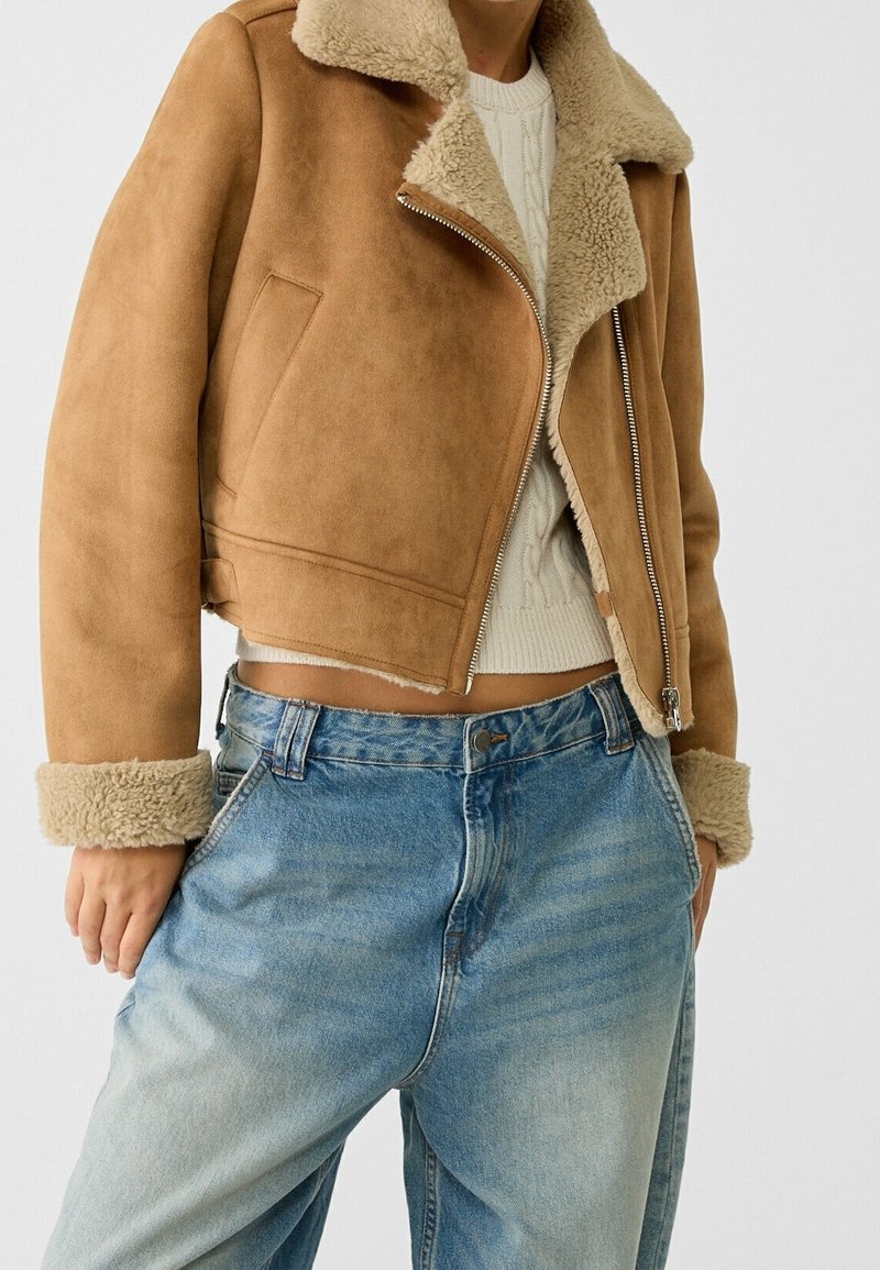 Krótka kurtka zamszowa w kolorze beżowym z miękkim beżowym kołnierzem i mankietami z owczej skóry, z zamkiem błyskawicznym i bocznymi kieszeniami. Noszona z jasnoniebieskimi dżinsami.