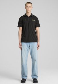 Sort polo shirt med standardkrave, prydet med grønne logoer og accenter. Bæres med blå denim jeans og sorte sko.