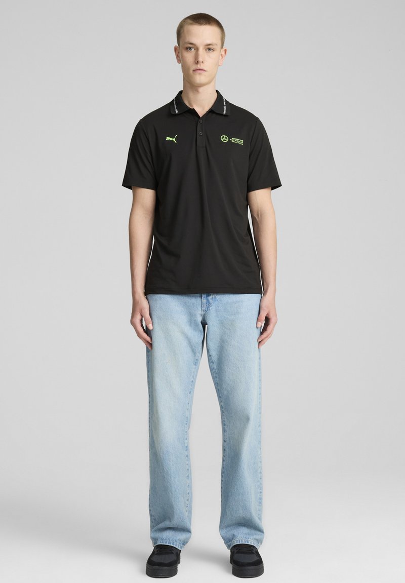 Sort polo shirt med standardkrave, prydet med grønne logoer og accenter. Bæres med blå denim jeans og sorte sko.