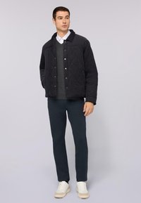 Chaqueta negra acolchada con cierres a presión sobre un suéter gris oscuro, combinada con pantalones azul marino y zapatillas claras. Tejido suave, corte moderno.