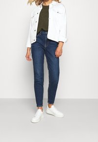 Weiße Jeansjacke über einem olivgrünen Strickpullover, kombiniert mit dunkelblauen Skinny Jeans und weißen Sneakers. Klare Linien und lässiges Design.