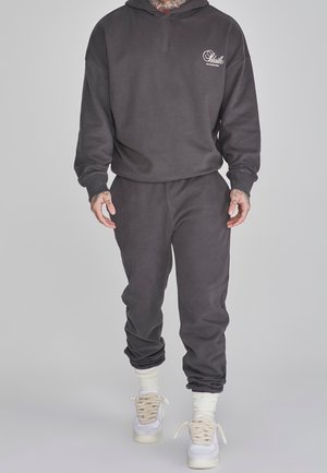 Mand iført mørkegrå hættetrøje og matchende joggingbukser, hvide sokker og hvide sneakers med tykke snørebånd, stående mod en ensfarvet baggrund.