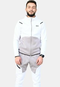 Chándal atlético blanco con acentos grises, que incluye una chaqueta con cremallera y pantalones ajustados, fabricado en un tejido suave; incluye detalles de herrajes en negro.