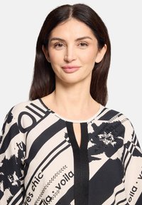 Sort og cremefarvet mønstret bluse med korte ærmer, med V-hals og central sort panel; indeholder tekst- og blomsterdetaljer.