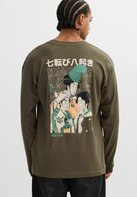 Camisa de manga larga verde oliva con un estampado gráfico de tres figuras con vestimenta tradicional, con texto en inglés y japonés.