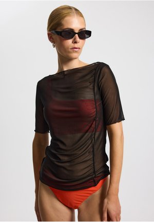 Schwarzes durchsichtiges Oberteil mit kurzen Ärmeln, mit gerafftem Design und rotem Akzent. Kombiniert mit orangefarbenen Bikinihosen.