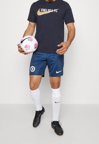 Camiseta de Nike del Chelsea FC en color marino con letras doradas, pantalones cortos azules con patrón de cuadrícula, calcetines blancos y botas de fútbol negras, sosteniendo un balón con patrón.