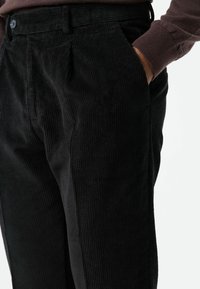 Zwarte corduroy broek met verticale ribbels, voorzien van zijzakken en een losse pasvorm. Tailleband met riemlussen en een knoopsluiting.