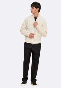 Cremefarbener Zip-Up-Pullover mit gerippten Ärmeln, getragen über einem schwarzen Hemd, kombiniert mit schwarzen Hosen und schwarzen Sneakers. Glatte Textur, taillierter Schnitt.