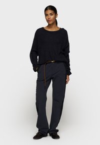 Donker trui en marineblauwe broek met een bruine riem. De trui heeft een losse pasvorm; de broek heeft zijpanelen en zakken.