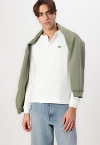 Weißes, geripptes Langarm-Polo mit einem grünen Pullover, der über die Schultern gelegt ist, kombiniert mit hellblauen Jeans.