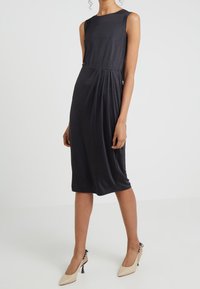 Vestido midi negro sin mangas con detalles plisados, textura suave y silueta ajustada. Combinado con zapatos de tacón alto color beige claro.