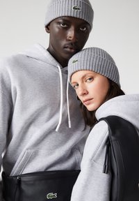 Deux personnes portant des bonnets gris assortis et des sweat-shirts à capuche avec un sac noir, regardant directement devant eux sur un fond uni.