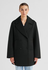 Schwarzer doppelt gefütterter Mantel mit breitem Revers, zwei Knöpfen und seitlichen Taschen; glatte Textur, übergroßer Schnitt und gerade Passform.