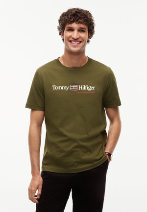 Smilende ung mand med krøllet hår iført olivengrøn Tommy Hilfiger t-shirt og mørke bukser, står med den ene hånd i lommen.
