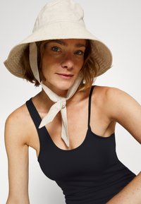 Chapéu de linho beje de aba larga com tiras amarradas, combinado com uma blusa preta de alças. O chapéu tem um acabamento texturado e um tom suave e natural.