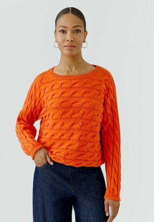 Frau trägt einen orangefarbenen Zopfpullover und dunkelblaue Jeans, steht mit einer Hand in der Tasche vor einem schlichten Hintergrund.