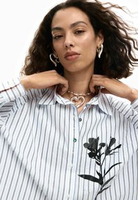 Sinine-valge triibuline nööbitav särk, mille vasakul küljel on must lillemuster ja hõbedane südamekujuline kaelakee. Lokkis juuksed ja kõrvarõngad.