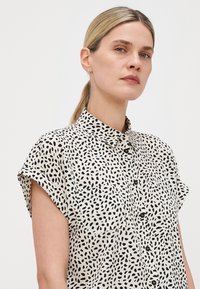 Chemise à manches courtes avec boutons en crème, ornée de taches abstraites noires, dotée d'un col, d'une poche poitrine et d'une coupe décontractée.