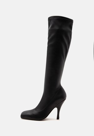 Botte en cuir noire montant jusqu'au genou, avec un bout pointu et un talon haut et fin sur fond blanc.