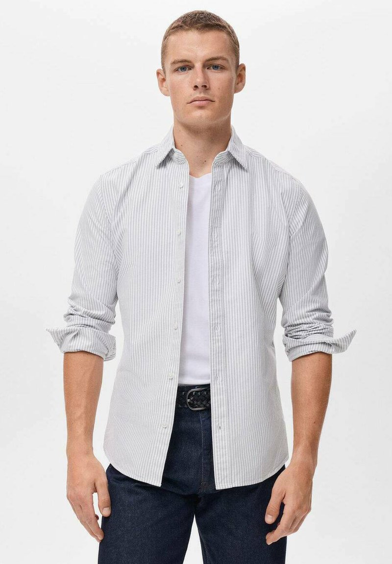 Camisa de botones a rayas en gris claro y blanco, de algodón, con bolsillos frontales y mangas enrolladas, llevada sobre una camiseta blanca, combinada con jeans oscuros.