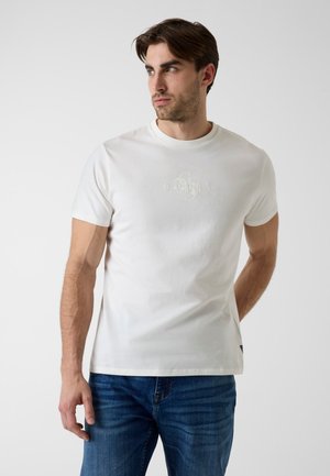 Homme portant un t-shirt blanc à manches courtes Guess et un jean bleu, debout légèrement tourné vers la gauche sur un fond clair et uni.