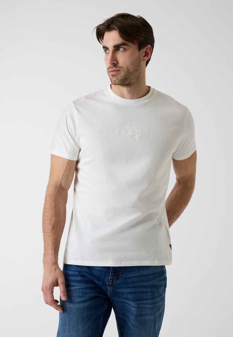Άνδρας με λευκό κοντομάνικο μπλουζάκι Guess και μπλε τζιν στέκεται ελαφρώς στραμμένος προς τα αριστερά μπροστά σε απλό ανοιχτόχρωμο φόντο.