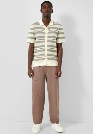Mężczyzna ubrany w beżowo-szarą koszulę na krótki rękaw w paski, brązowe spodnie o szerokich nogawkach i białe trampki stoi na jednolitym tle.