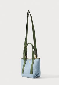 Τσάντα tote από ανοιχτό μπλε ύφασμα με σκούρες πράσινες λαβές, χρυσά μεταλλικά στοιχεία και ρυθμιζόμενο μακρύ λουράκι ώμου, πάνω σε απλό λευκό φόντο.