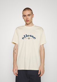 Camiseta de algodón crema con mangas cortas y escote redondo. Presenta el logo y texto de "ellesse" en azul marino con un diseño curvado.