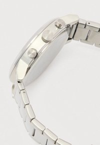 Sudraba metāla rokas pulkstenis ar saistītām siksnām, divām spiedpoga un teksturētu vainagu malā, attēlots no sānu leņķa.