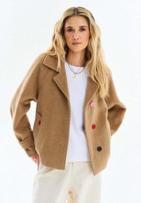 Žena oblečená v béžovom vlnenom saku s farebnými gombíkmi cez bielu košeľu a krémové nohavice, stojaca na jednoduchom bielom pozadí.