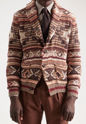 Mann trägt braunen gemusterten Strickcardigan über beige Hemd und dunkelbraune Krawatte, kombiniert mit brauner Hose.
