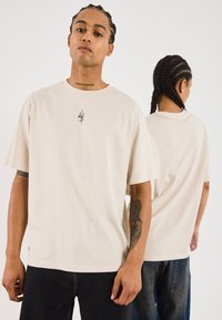 Beige Baumwoll-T-Shirt mit kurzen Ärmeln, das ein kleines schwarzes Grafikdesign auf der Brust präsentiert. Lässige Passform und minimalistisches Design.