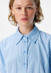 Chemise rayée bleu clair avec un col pointu et des manches courtes, dotée de boutons et d'une texture de tissu douce.