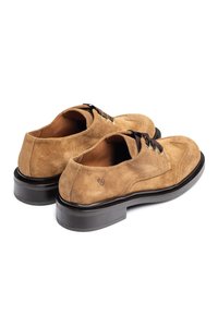 Chaussures en suède marron avec une texture lisse, dotées de coutures courbes, de lacets noirs et d'une semelle noire épaisse. Design simple avec des accents minimaux.